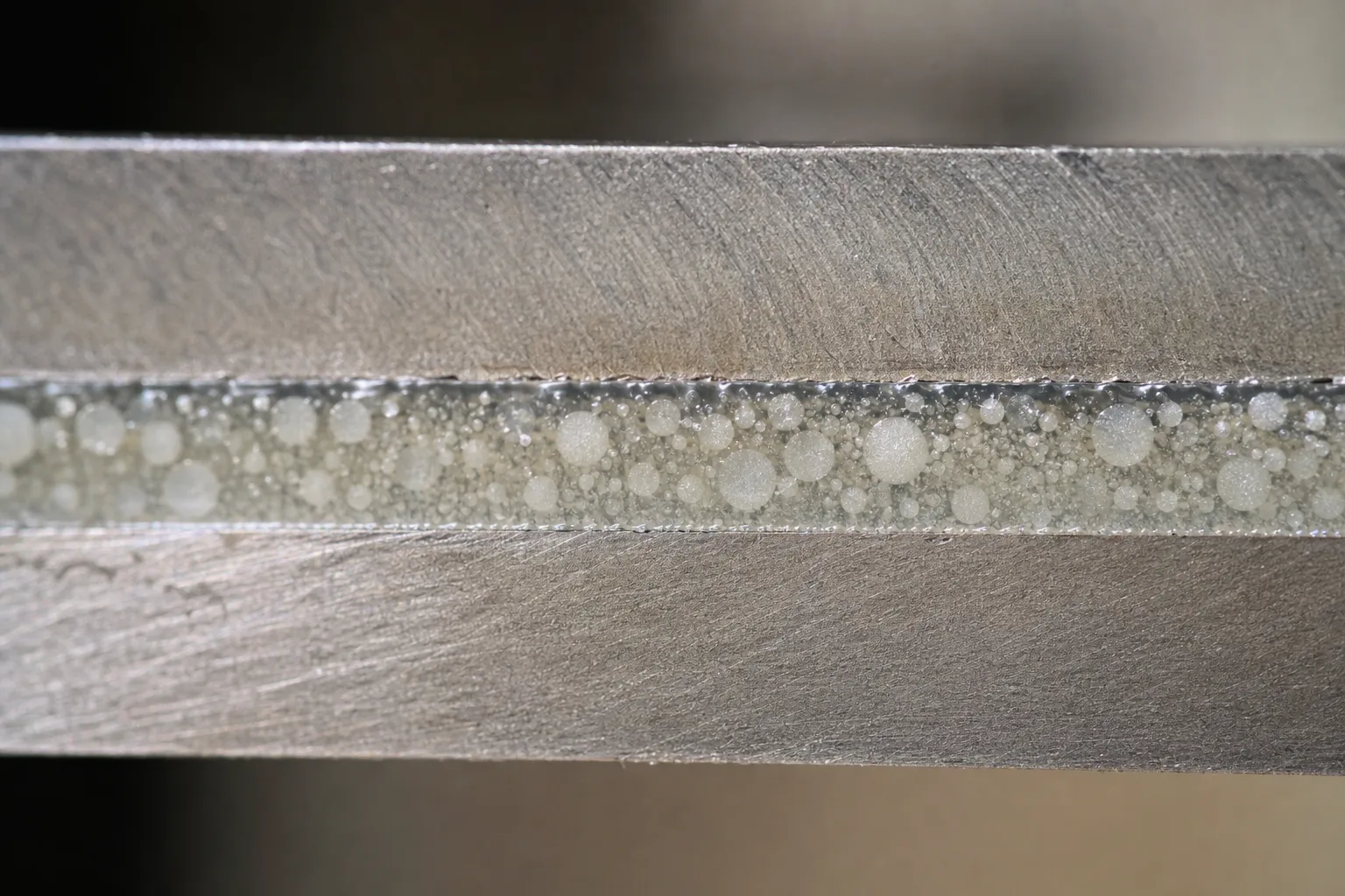A detailed view of a rubber toughened cyanoacrylate adhesive joint between two metal parts in a controlled manufacturing environment. The image shows the microstructure contributing to shock and heat resistance.
